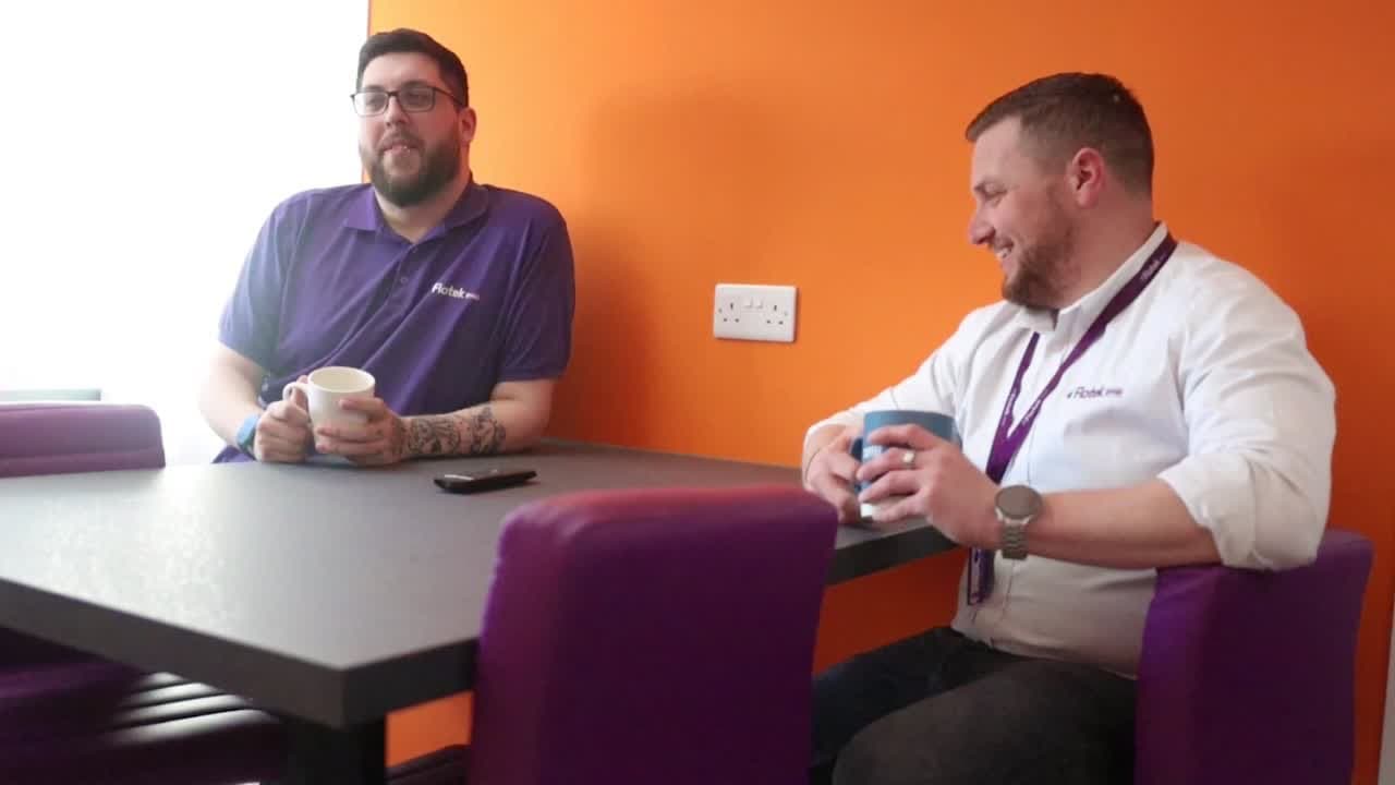 Two members of Flotek's team in purple Flotek shirts talking over coffee at a table in front of an orange wall.