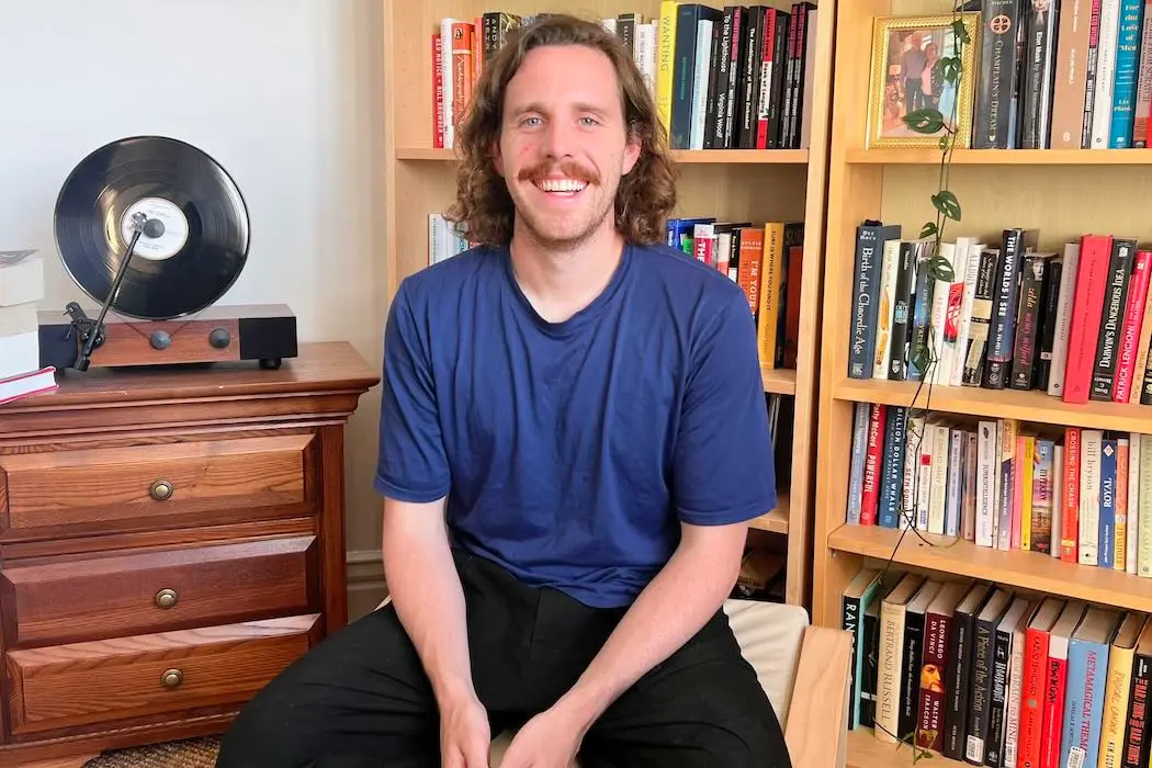 Marc-André Campagna, gaiia's co-founder and CEO, seated and smiling in front of a full bookshelf, with a record player on a dresser beside him.